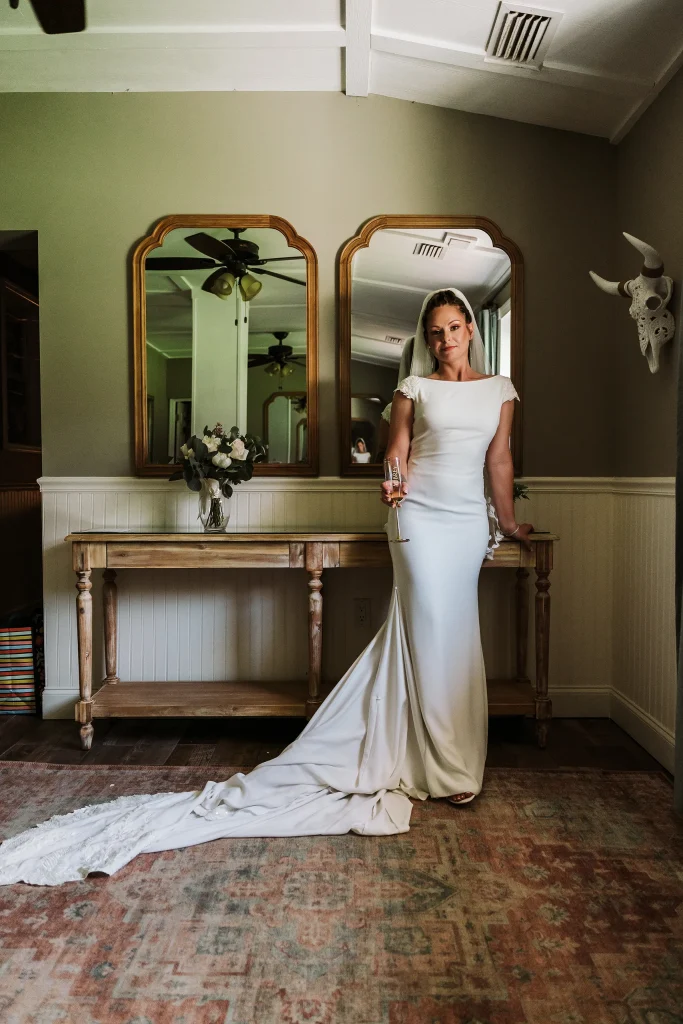 Bride standing in the Masters Stables bridal suite wearing a fitted wedding gown with a long train, holding a champagne glass in an elegant indoor portrait.