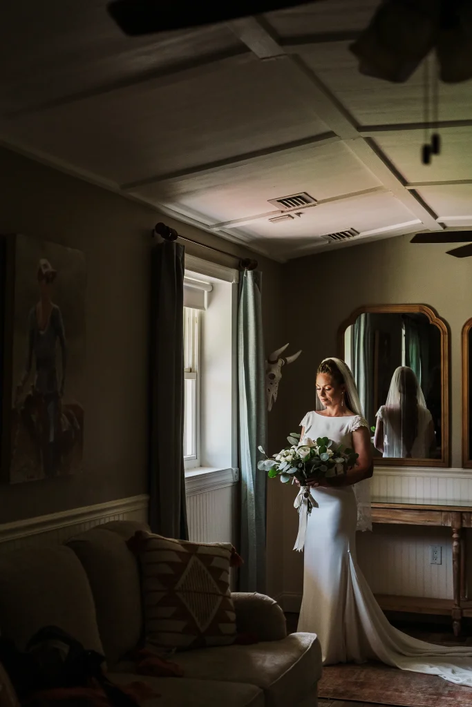 Bride standing in soft window light inside the Masters Stables bridal suite, holding a greenery bouquet in an elegant fitted wedding gown.