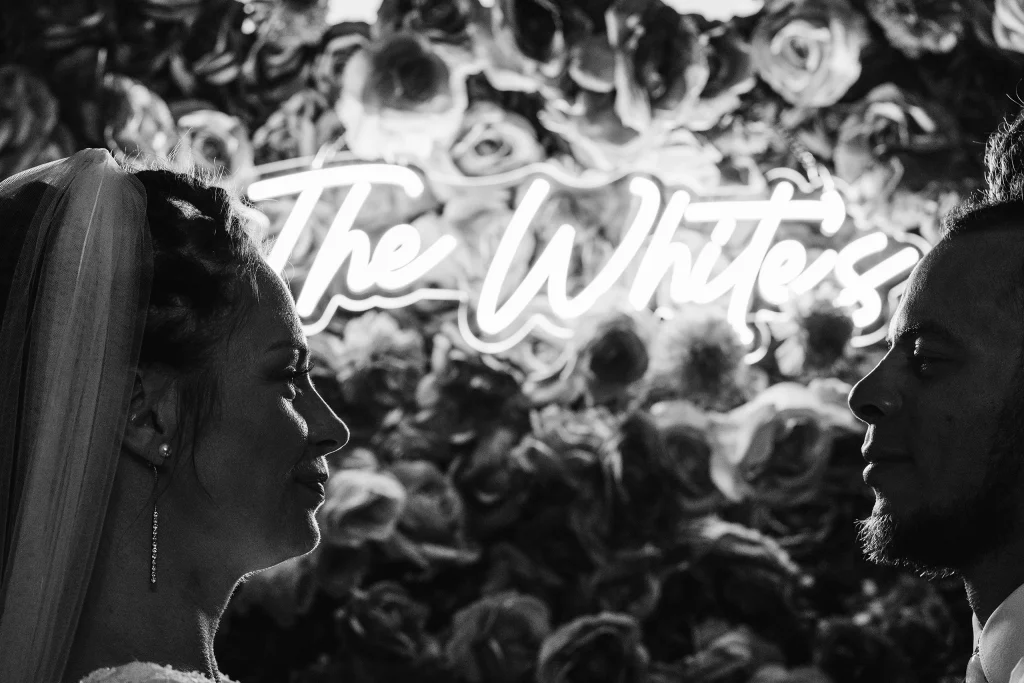 Bride and groom face each other in silhouette in front of their custom neon &ldquo;The White&rsquo;s&rdquo; sign and floral backdrop during their Masters Stables wedding.