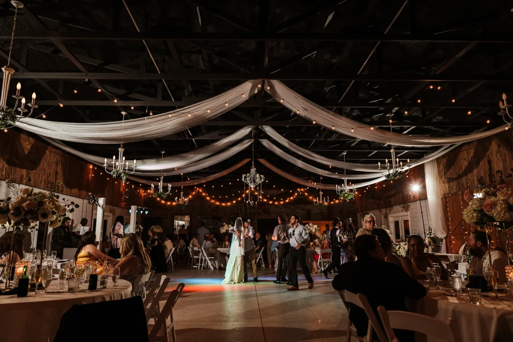 Guests dancing during the evening wedding reception at Masters Stables in Fellsmere, Florida, with draped ceilings, chandeliers, string lights, and decorated tables surrounding the dance floor.